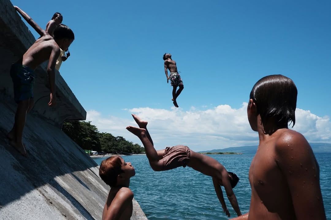 Artist Spotlight: “Catch A Moment” by @hersleyvencasero

This weeks Artist Spotlight features Hersley-Ven Casero, multidisciplinary visual artist and photographer based in Dumaguete City, Philippines. His long-term series, “Catch A Moment” moves through everyday streets and fleeting glances, finding honesty in the uncanny moments that make us deeply human.

“These photographs are not about perfection. They are about presence. I’m chasing a feeling: the rush of kids mid-air above the sea, the hush of light falling through a window, a dog silhouetted against a golden sky, kids playing atop sacks of crimson onions, glowing like jewels, a woman’s face caught twice in one moment, real and reflected. Every frame is a quiet witnessing, a heartbeat stilled in time.

Drawn to the poetry of the everyday, I look for the magic in the mundane: in the warmth of sunlit walls, in shadows cast by strangers, in the choreography of street life unfolding without warning. This is where stories live, where life pulses loudest in its smallest, most honest gestures.

Catch A Moment is my way of honoring what most might walk past. It is not about capturing the perfect shot. It is about capturing the true one, the kind that stirs something in you, the kind you feel before you fully see.”